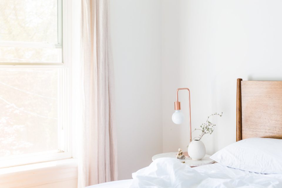 Bright bedroom with a window, white bedding and a bedside lamp in the morning sun