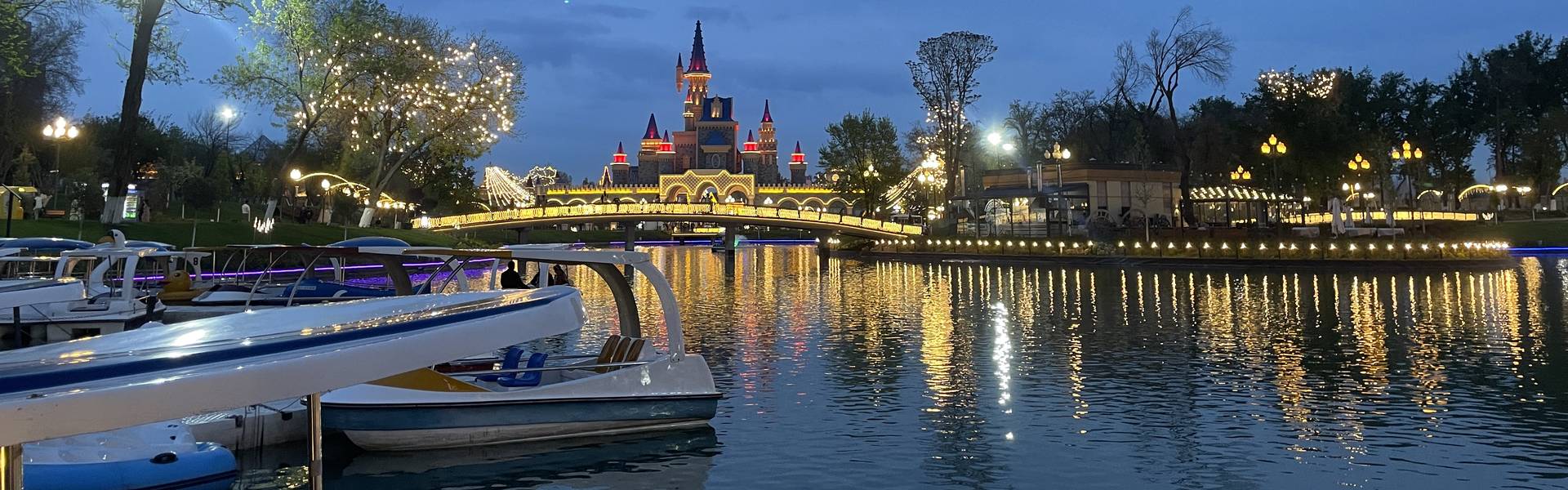 Замок парка Magic City вечером над озером в Ташкенте, мост с огнями и катамараны у причала