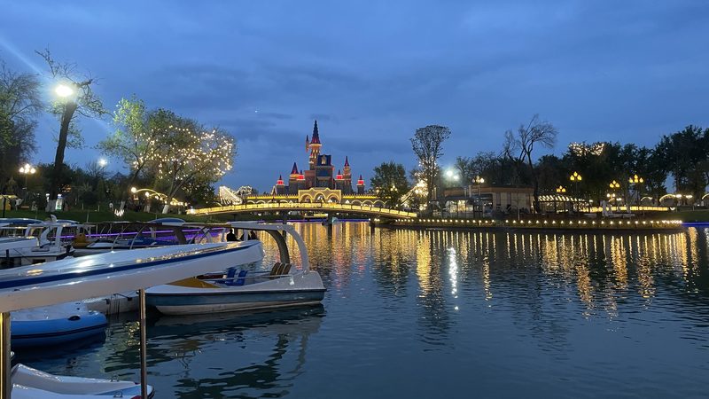 Замок парка Magic City вечером над озером в Ташкенте, мост с огнями и катамараны у причала